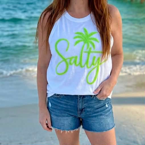 Salty Fluffy Patch, sleeveless white tank top with green palm tree and stylish script logo, worn by a woman on the beach.