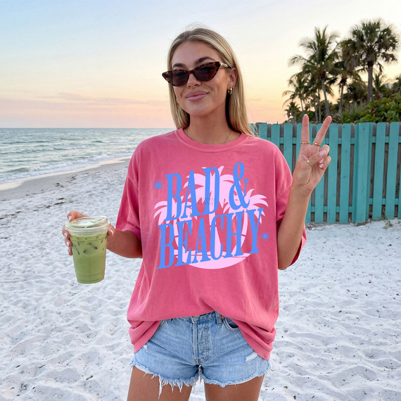 Bad & Beachy DTF Heat Transfer, a pink T-shirt with a colorful beach-themed design, worn by a woman on the beach during sunset.