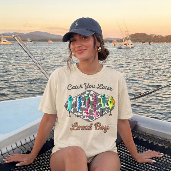 Catch Ya Later Local Boy, image of a woman on a boat wearing a T-shirt with a fishing lure design and the words 'Catch Ya Later Local Boy', sunset and boats in the background.