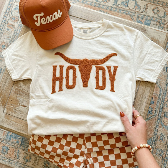 Howdy Longhorn Fluffy Patch, close-up of a white T-shirt with a textured orange longhorn patch and letters, paired with a brown and white checkered skirt and a rust-colored cap with 'Texas' embroidered on it.