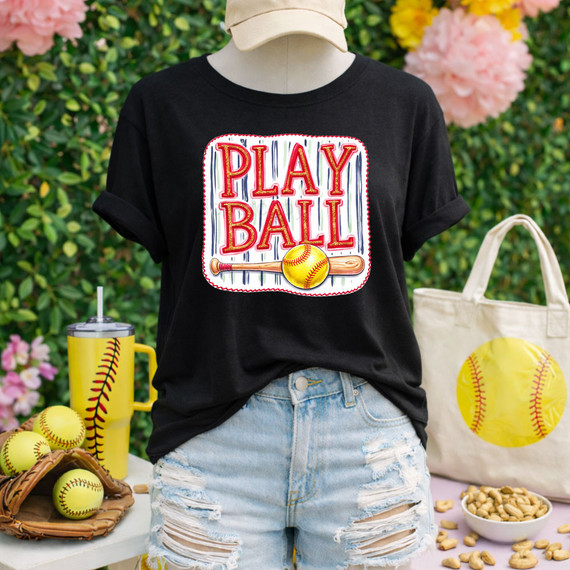 Play Ball Softball DTF Heat Transfer, colorful graphic design with a baseball and bat on a black T-shirt worn by a mannequin outdoors surrounded by baseball-themed accessories.