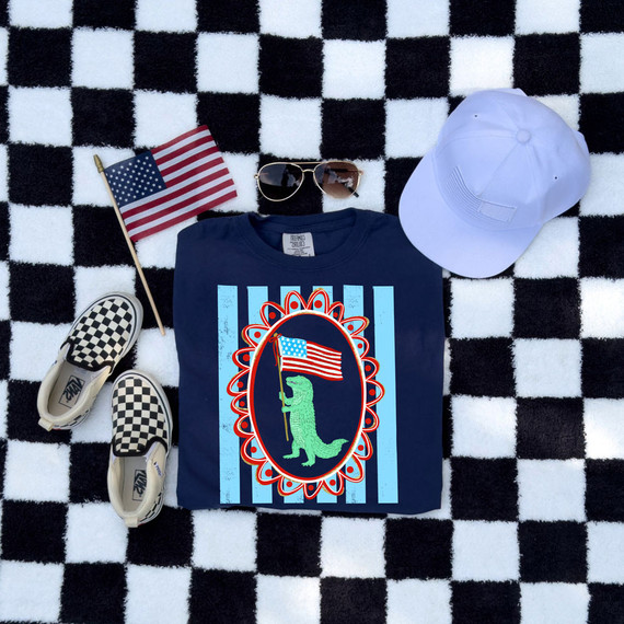Patriotic Dino Flag DTF Heat Transfer, patriotic-themed t-shirt with a dinosaur holding an American flag, placed on a black and white checkered background, accompanied by sunglasses, a white cap, a small American flag, and checkered footwear.