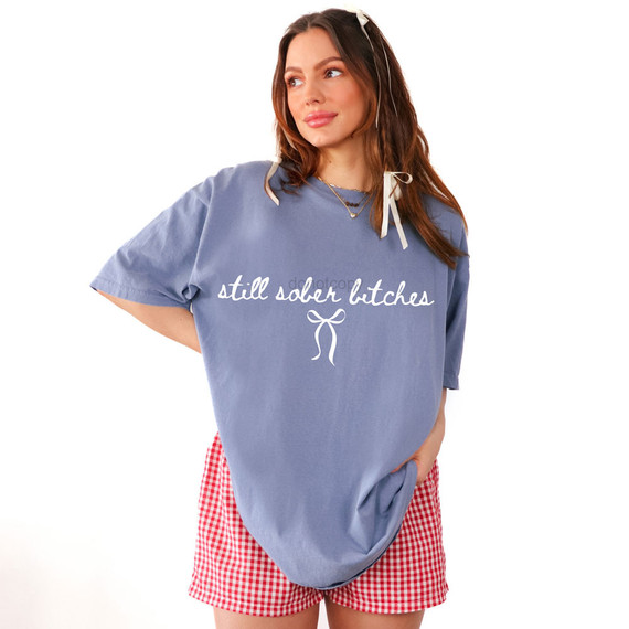 Still Sober Bitches WHITE DTF Heat Transfer, white text design on a blue T-shirt worn by a woman in casual red and white checkered shorts.