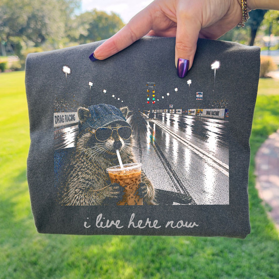 I Live Here Now Drag Race DTF Heat Transfer, product image showing a dark grey clothing item with a graphic of a raccoon wearing sunglasses and sipping an iced drink, set against a backdrop of a drag racing track.