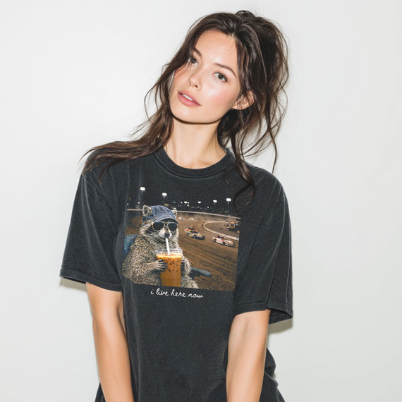 i Live Here Now Dirt Track DTF Heat Transfer, a woman wearing a black t-shirt with a graphic of a raccoon drinking bubble tea at a dirt track racing event.