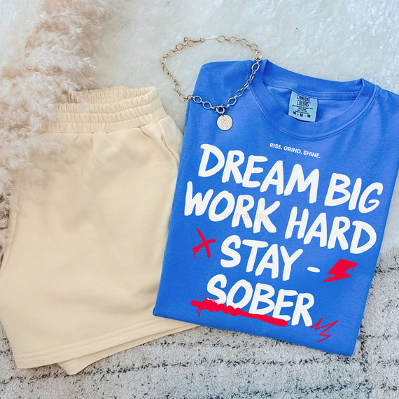 Dream Big Work Hard Stay Sober DTF Heat Transfer, blue t-shirt with bold white text and red graphic accents, paired with cream-colored shorts and a silver chain necklace.