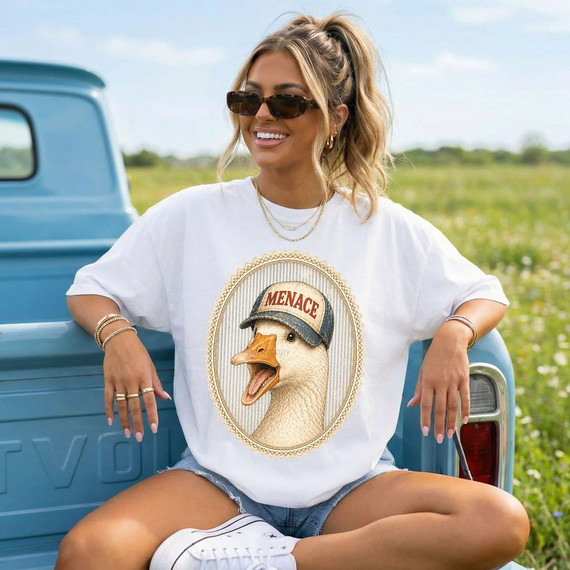 Menace Goose DTF Heat Transfer, product image of a woman wearing a white t-shirt with a cartoon goose wearing a denim cap with 'MENACE' written on it, showcased outdoors sitting on a vintage blue truck bed.