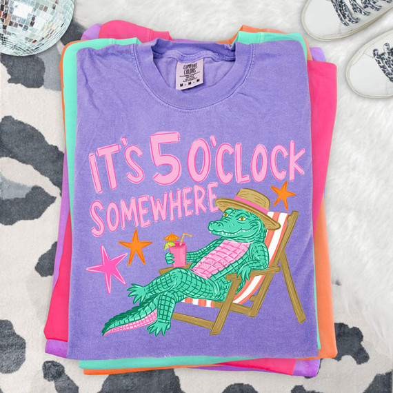 It's 5 O'Clock Somewhere Gator DTF Heat Transfer, colorful graphic T-shirt featuring an alligator relaxing with a drink, in a lounging chair, wearing a sun hat on a folded pile of clothing.