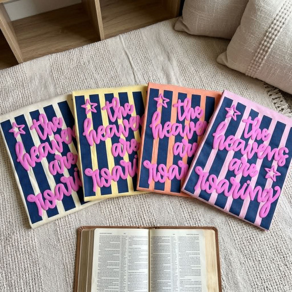 The Heavens Are Roaring Puff Screen Print Transfer, colorful fabric transfer with the phrase 'The Heavens Are Roaring' in pink lettering and star accents, laid out on a beige textured surface.
