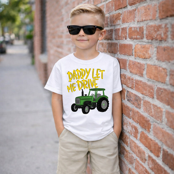 Daddy Let Me Drive Tractor DTF Heat Transfer, a young boy wearing sunglasses and a white T-shirt with a green tractor and yellow text printed on it, standing outdoors against a brick wall.