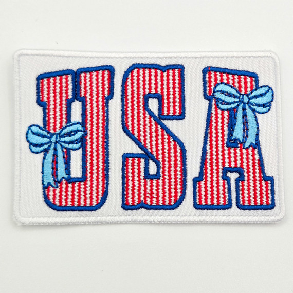 USA Coquette Embroidered Hat Patch – 3.5” Wide, embroidered patch with red and white striped letters 'USA' outlined in blue, featuring two light blue bows on the 'U' and 'A', designed to be sewn onto hats or clothing.
