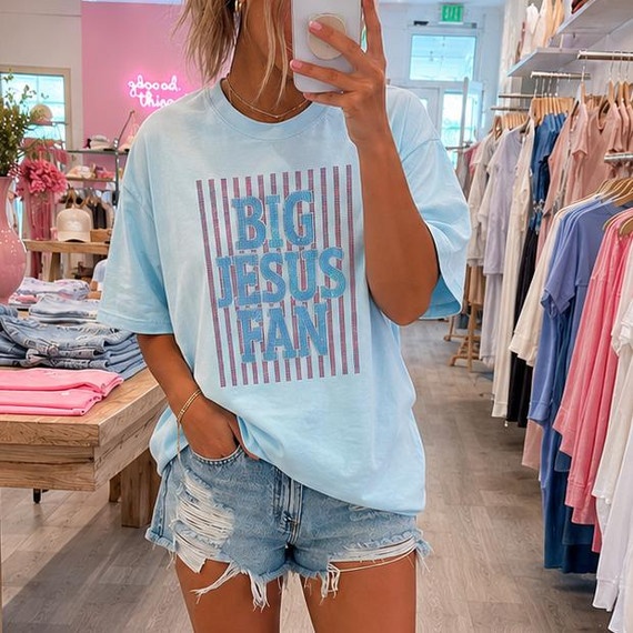 Big Jesus Fan Spangle Transfer, a woman wearing a light blue t-shirt with a glittery 'Big Jesus Fan' design and distressed denim shorts, taking a mirror selfie inside a clothing boutique.