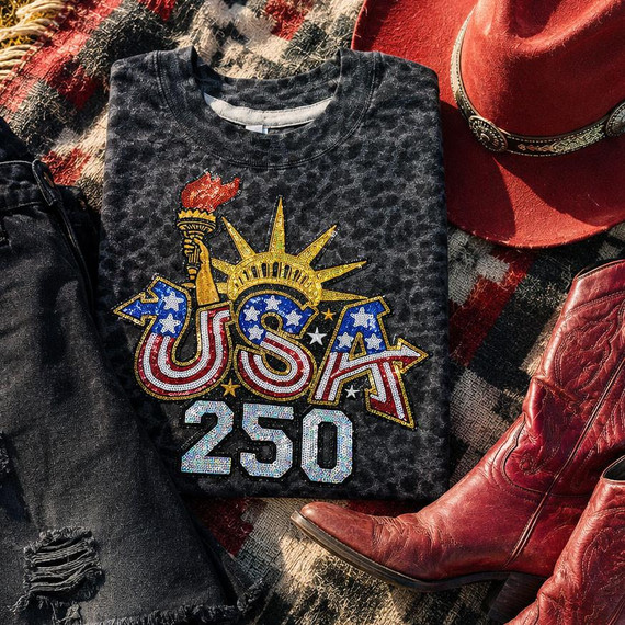 USA 250 Sequin Patch, patriotic embroidered patch with American flag colors, torch, and star design on a black textured T-shirt, surrounded by red cowboy boots, a red cowboy hat, and black distressed jeans on a checkered fabric background.
