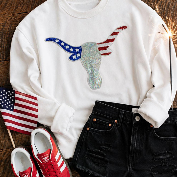 Patriotic Longhorn Sequin Patch, close-up of a sweatshirt with a sparkly sequin patch of a longhorn with American flag motifs on the horns, surrounded by American flags and red and white sneakers on a wooden surface.
