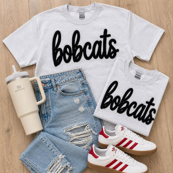 Bobcats Fluffy Chenille Patch Transfer, varsity spirit wear patch on grey t-shirts, casual denim shorts, white and red Adidas sneakers, and a beige Stanley water bottle laid out on a wooden surface.