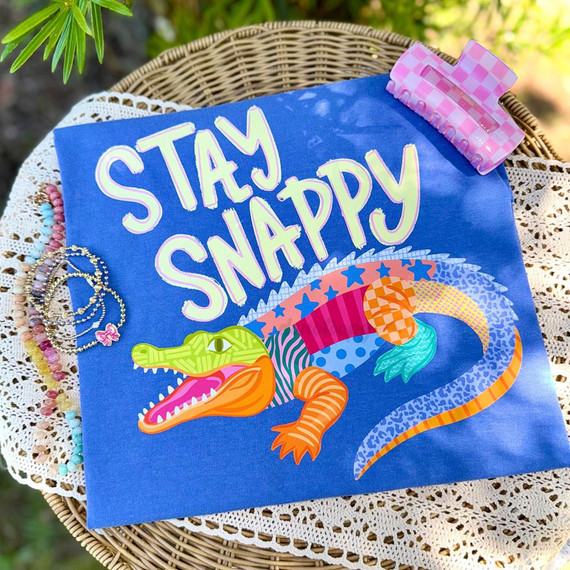 Stay Snappy DTF Heat Transfer, colorful animated crocodile with vibrant patterns and the phrase 'Stay Snappy' on a blue fabric background.