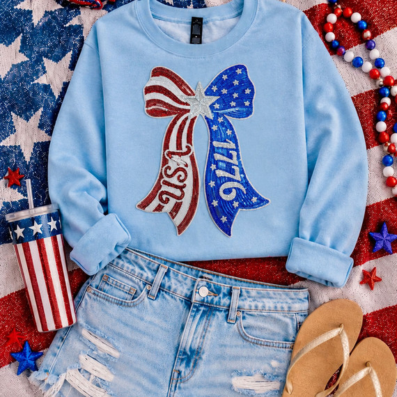 Light blue patriotic sweatshirt featuring a large red, white, and blue 1776 USA sequin bow patch styled with distressed denim shorts, flip flops, and Fourth of July accessories.