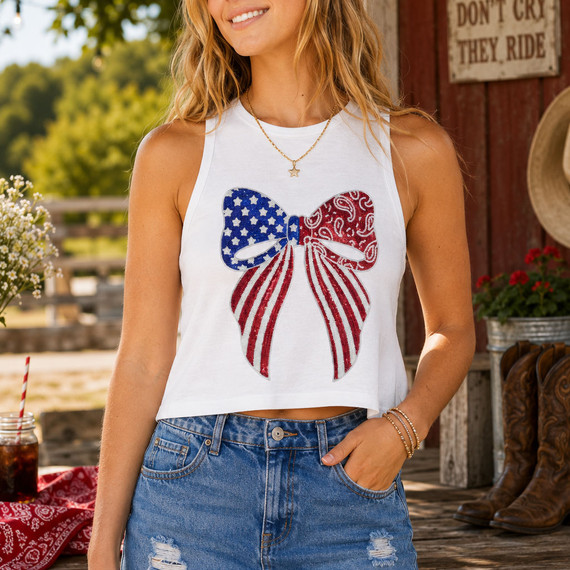 Woman wearing a white tank top featuring a large patriotic red, white, and blue sequin bow patch styled with denim shorts and western Americana accessories.