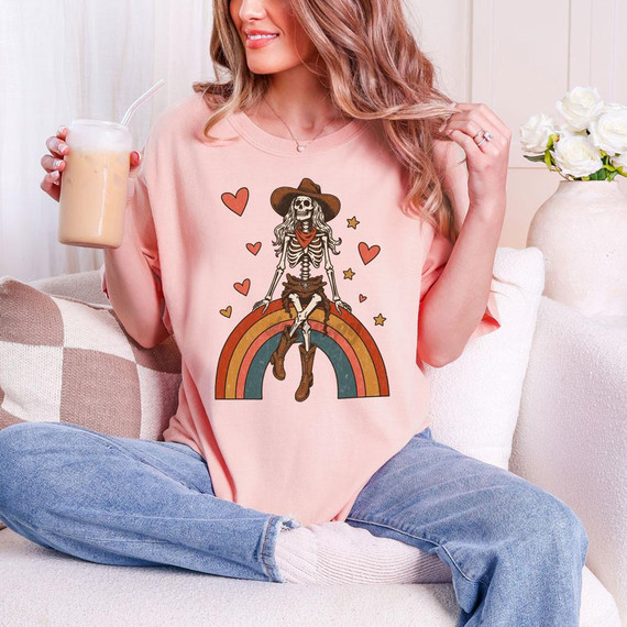 Cowgirl Skeleton Rainbow DTF Heat Transfer, colorful skeleton design on a pink T-shirt with rainbow, hearts, stars, and cowgirl hat features.
