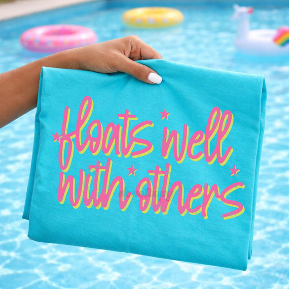 Floats Well With Others DTF Heat Transfer, image of a colorful towel with the phrase "Floats Well With Others" in pink and yellow lettering, held by a hand against a pool with inflatable floats.