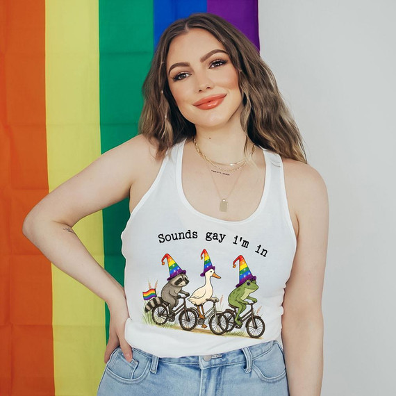 Sounds Gay I'm In DTF Heat Transfer, woman wearing a sleeveless white tank top with colorful cartoon animals riding bikes with rainbow hats and pride flags in the background.
