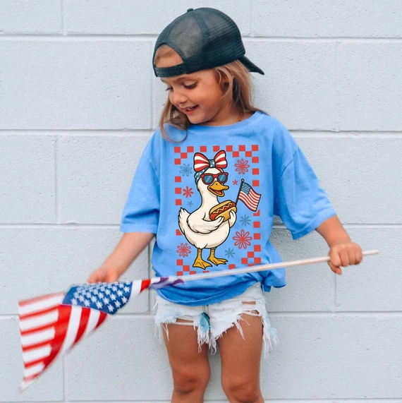 Patriotic Bow Duck DTF Heat Transfer, a girl wearing a patriotic-themed t-shirt with a cartoon duck holding an American flag and hot dog, celebrating Independence Day.