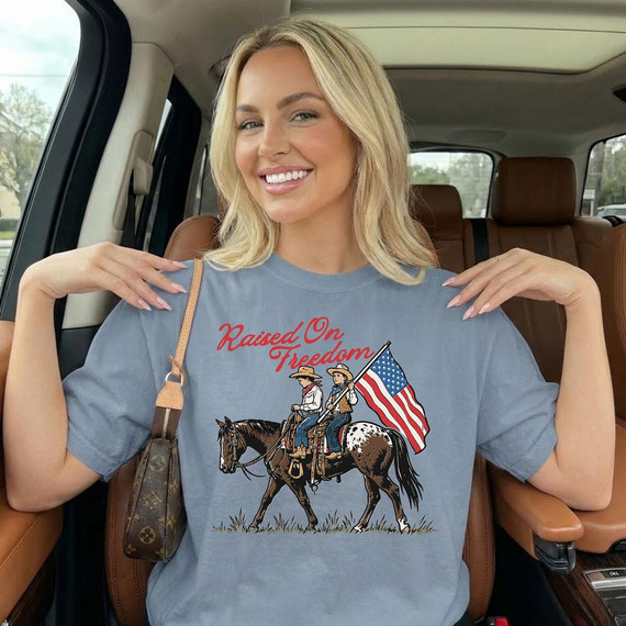 Raised On Freedom DTF Heat Transfer, woman wearing a t-shirt with a patriotic design of two riders on horseback with an American flag and the text 'Raised On Freedom'.