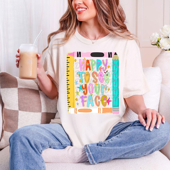 Happy To See Your Face Pencil Boarder DTF Heat Transfer, colorful cartoon-style design on a white t-shirt worn by a woman sitting on a sofa with a drink in her hand.