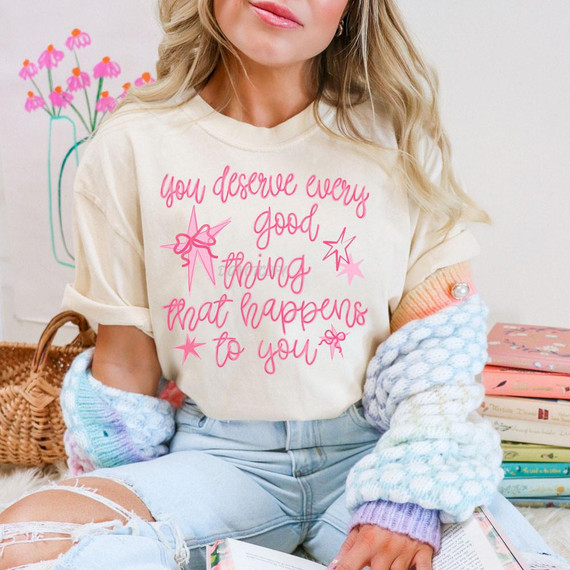 You Deserve Every Good Thing That Happens To You DTF Heat Transfer, a motivational pink text design on a cream-colored t-shirt worn by a woman sitting in a cozy, colorful setting.