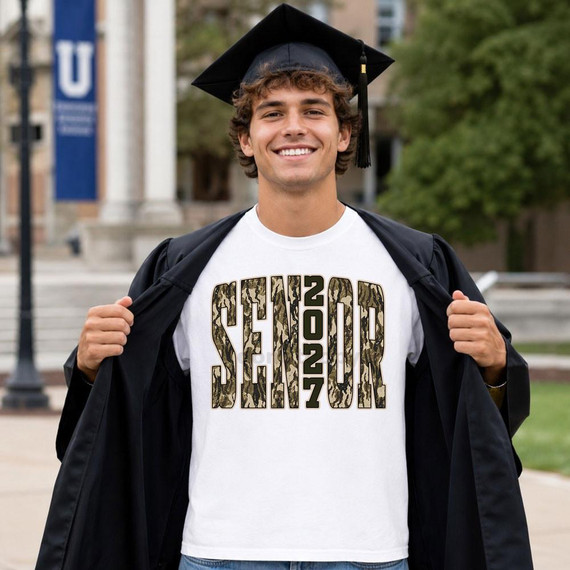 Senior 2027 Camo DTF Heat Transfer, a close-up of a young man in graduation attire showing the printed design on his white T-shirt featuring the text 'SENIOR 2027' in camouflage pattern.
