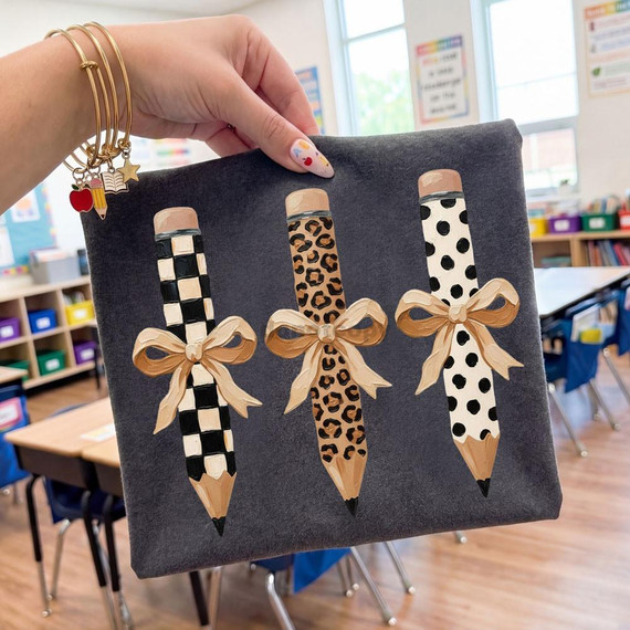 Coquette Pencil Trio DTF Heat Transfer, decorative pencils with leopard, checkered, and polka dot patterns with gold bows on a dark fabric canvas, classroom background.