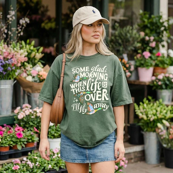 Some Glad Morning CREAM DTF Heat Transfer, woman wearing a green T-shirt with a vivid printed design and quote, standing among colorful flowers.