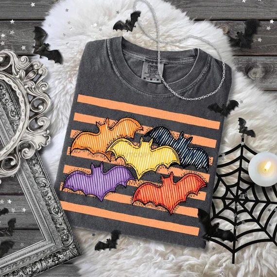 Colorful Bats DTF Heat Transfer, image of a T-shirt with colorful bat designs in orange, yellow, black, purple, and red, set against orange and black striped background with Halloween themed decorations.