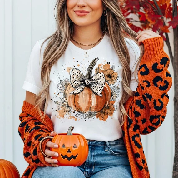 Polka Dot Bow Pumpkin DTF Heat Transfer, woman wearing fall-themed t-shirt with pumpkin and bow design, holding a pumpkin mug, autumn leaves background.