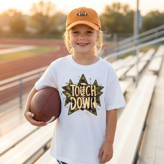 Touch Down Camo Stars DTF Heat Transfer, a girl standing on a sports track wearing a white T-shirt with a camo star design and text, holding a football.