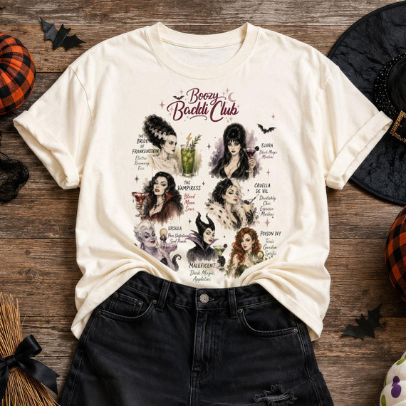 Boozy Baddie Club Halloween DTF Transfer, illustrated graphic t-shirt featuring fictional characters with Halloween themed names and designs, surrounded by Halloween decorations on a wooden surface.