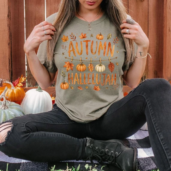 Autumn Hallelujah DTF Heat Transfer, a woman wearing an autumn-themed t-shirt with pumpkins and leaves design, sitting outdoors among pumpkins and fall decorations.