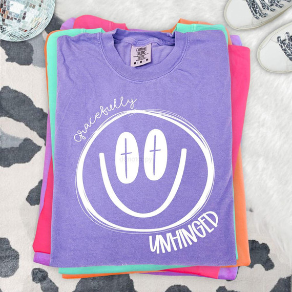 Gracefully Unhinged DTF Heat Transfer, vibrant purple T-shirt with a white smiley face and playful text design.