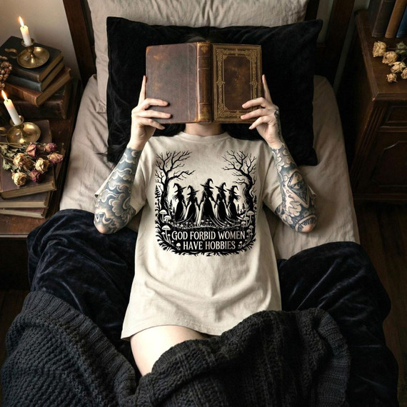 God Forbid Women Have Hobbies Halloween DTF Heat Transfer, woman lying in bed with a book covering her face, wearing a Halloween-themed T-shirt with witch silhouettes and trees.