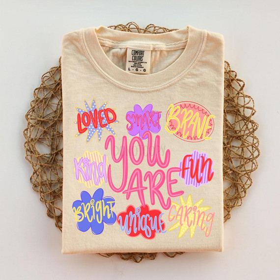You Are Loved Smart Brave Kind Fun Bright Unique Caring DTF Heat Transfer, colorful design on a folded beige T-shirt.