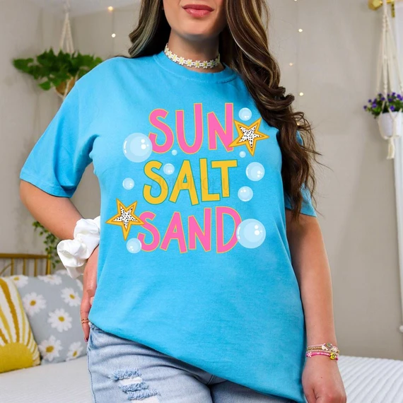 Colorful Sun Salt Sand DTF Heat Transfer, vibrant t-shirt design featuring bright pink, yellow, and white text with star and bubble graphics on a blue shirt.