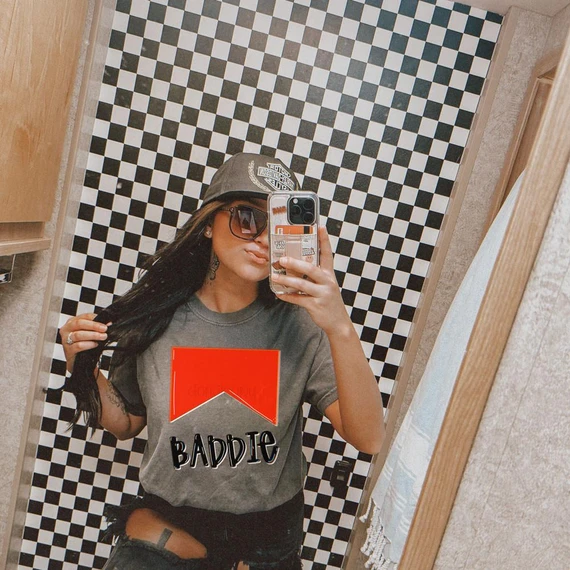 Grunge Baddie DTF Heat Transfer, trendy woman taking a selfie in a checkered bathroom mirror wearing a gray T-shirt with the Baddie graphic design.