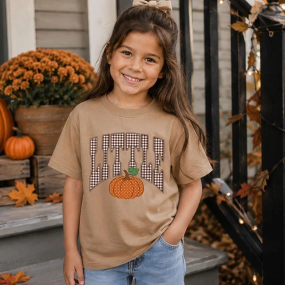 Gingham Fall Little DTF Heat Transfer, girl wearing a beige T-shirt with autumn-themed pumpkin and gingham design, outdoors with fall decorations.