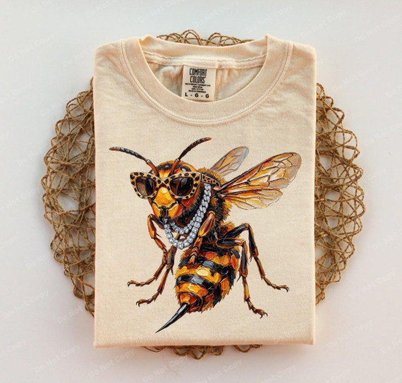 Glam Hornet DTF Heat Transfer, detailed printed design of a bee wearing sunglasses and a pearl necklace on a folded beige T-shirt.