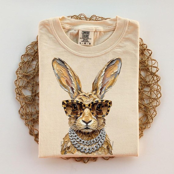 Glam Jackrabbit DTF Heat Transfer, beige T-shirt with a rabbit wearing sunglasses and a chain, laid on a woven round mat