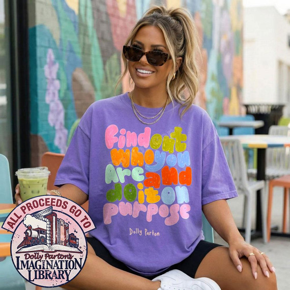 Find Out Who You Are And Do It On Purpose DTF Transfer - Dolly Parton Quote Design on purple t-shirt worn by a smiling woman with sunglasses, sitting outdoors with colorful mural background.