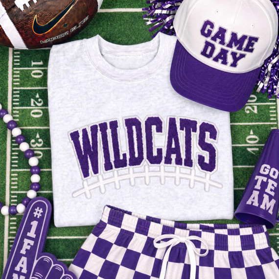 Wildcats Purple & White Chenille Football Patch, close-up view of the patch on a white shirt with purple lettering, surrounded by sports-themed accessories on a green field background.