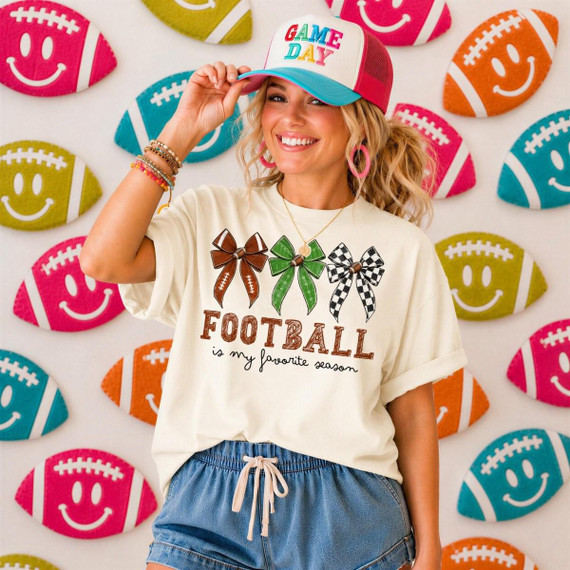 Football Is My Favorite Season DTF Transfer product, a woman wearing a white t-shirt with football bows and text, and a colorful 'Game Day' cap, standing in front of a backdrop decorated with football emojis.
