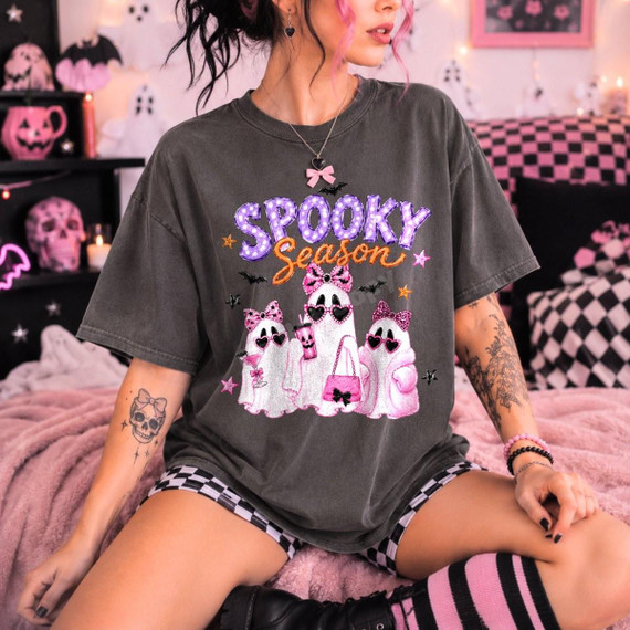 Spooky Season Girly Ghosts DTF Transfer, pink Halloween design on a dark gray t-shirt featuring cute ghost illustrations with pink bows and sunglasses, surrounded by stars and bats, worn by a woman in a festive Halloween room.