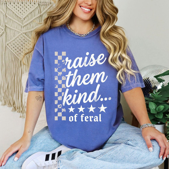 Raise Them Kind... Of Feral DTF transfer in distressed white script with a checkered stripe accent and stars on a periwinkle shirt